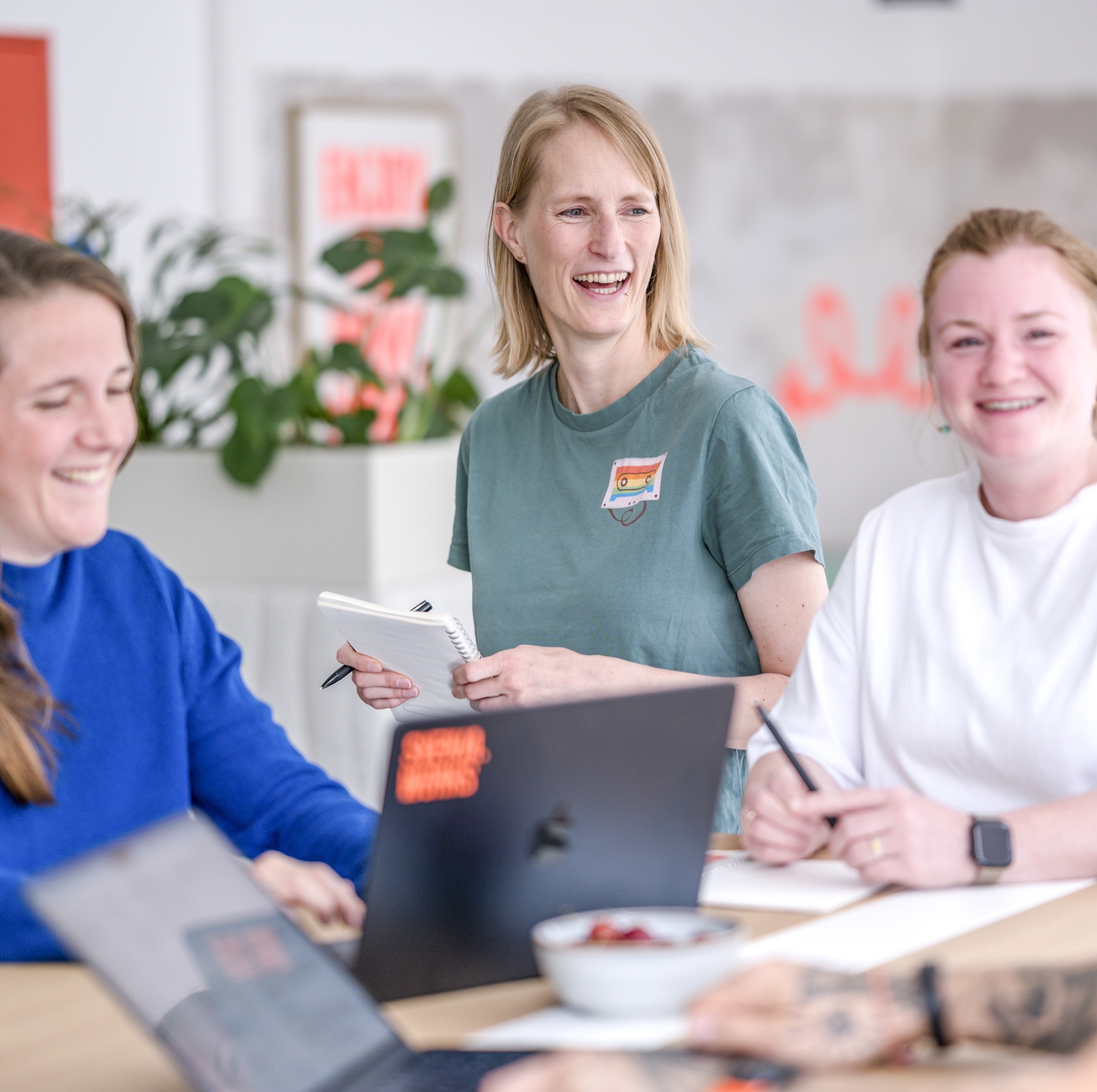Drei lachende Frauen mit Laptops und Notizbüchern an einem Tisch im glæd.works-Büro während eines Team-Meetings.