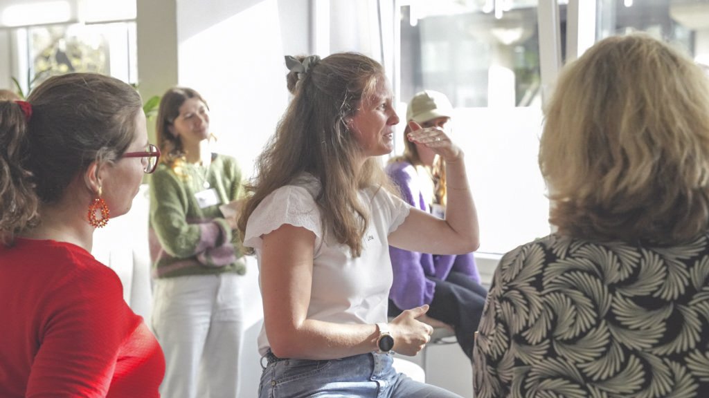 Frau im weißen Shirt gestikuliert im Gespräch innerhalb einer Gruppe in einem hellen, sonnendurchfluteten Raum.
