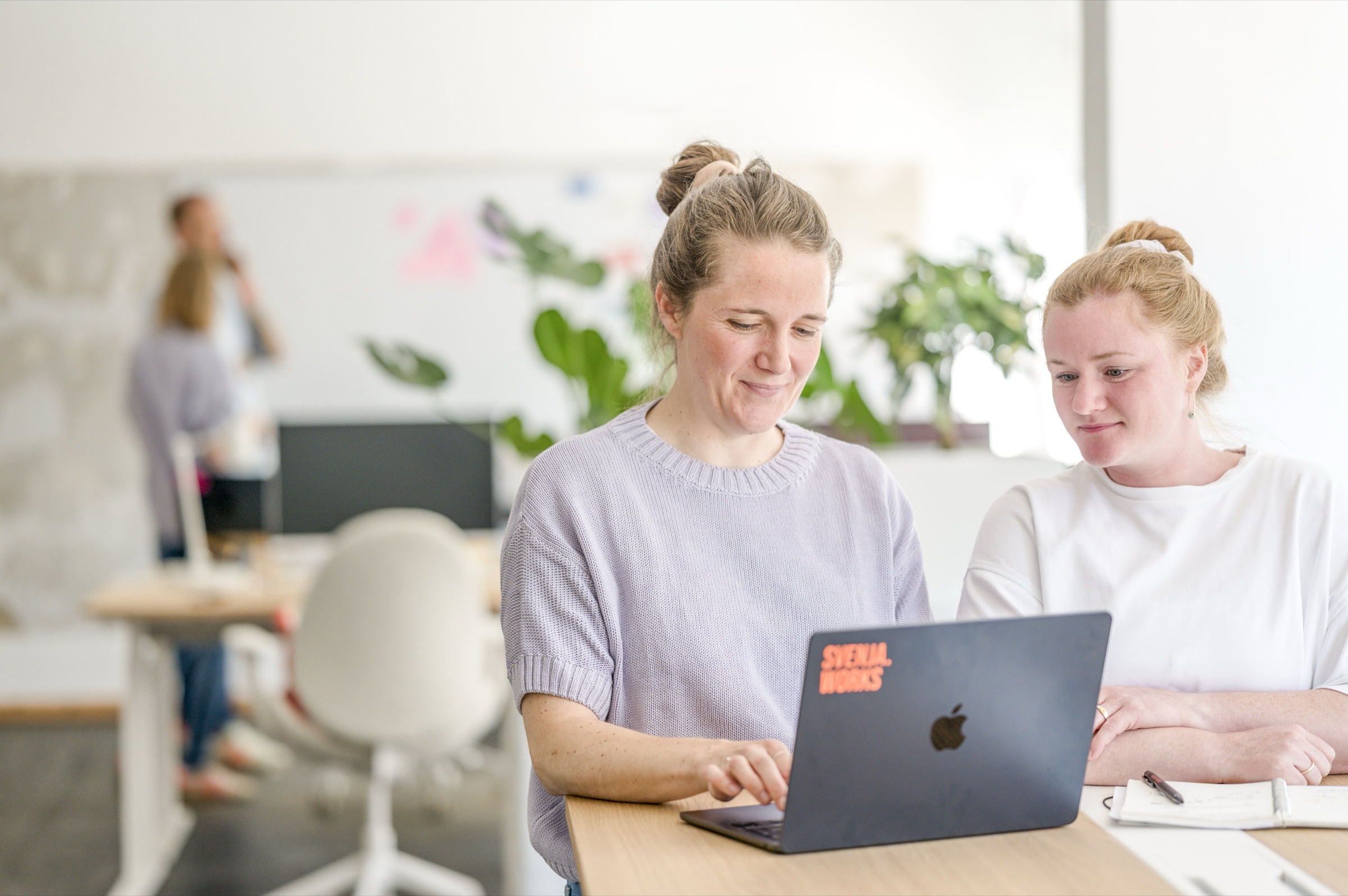 Zwei Frauen schauen lächelnd gemeinsam auf einen Laptop-Bildschirm in einem Büro mit Pflanzen.
